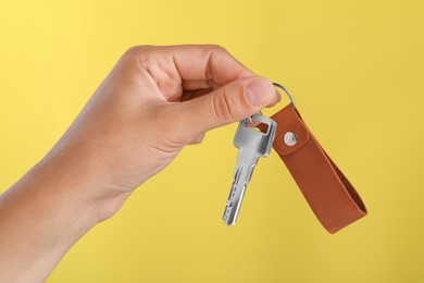 Photo of Woman holding key with leather keychain on yellow background, closeup