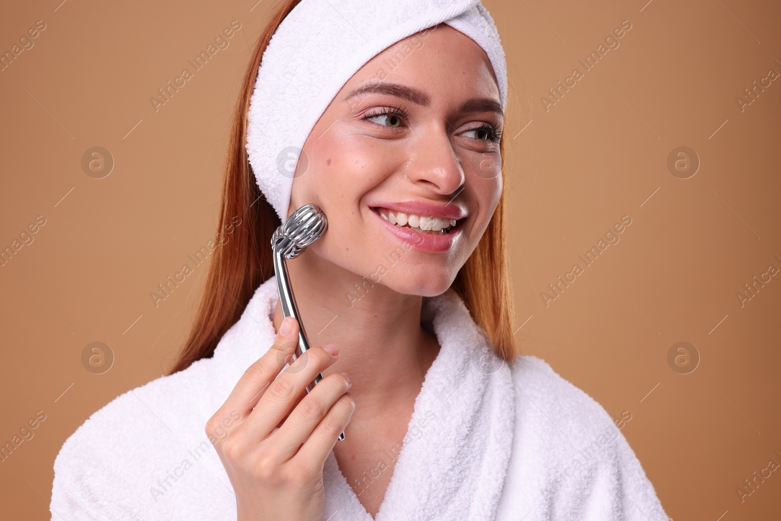 Photo of Young woman massaging her face with metal roller on pale orange background