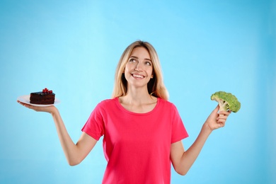 Woman choosing between cake and healthy broccoli on light blue background