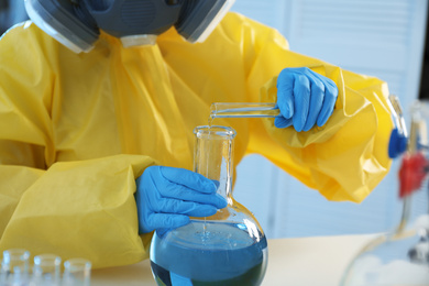 Scientist in chemical protective suit pouring reagent into flask at laboratory, closeup. Virus research