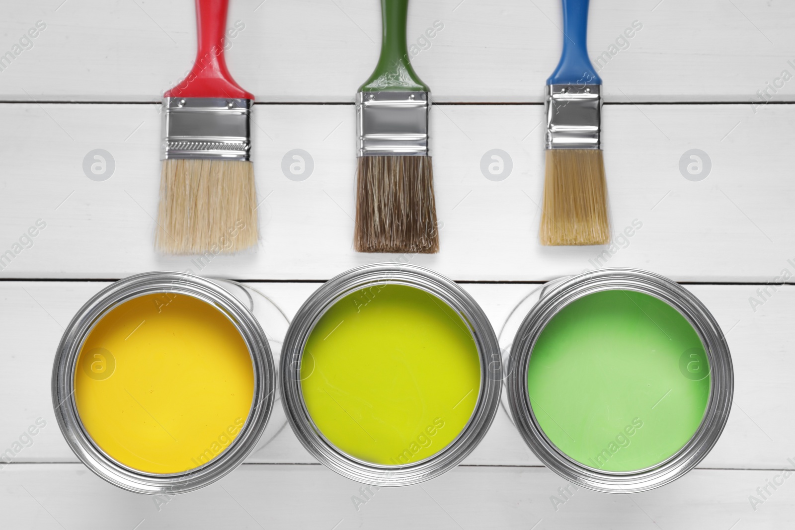 Photo of Cans of yellow and green paints with brushes on white wooden table, flat lay