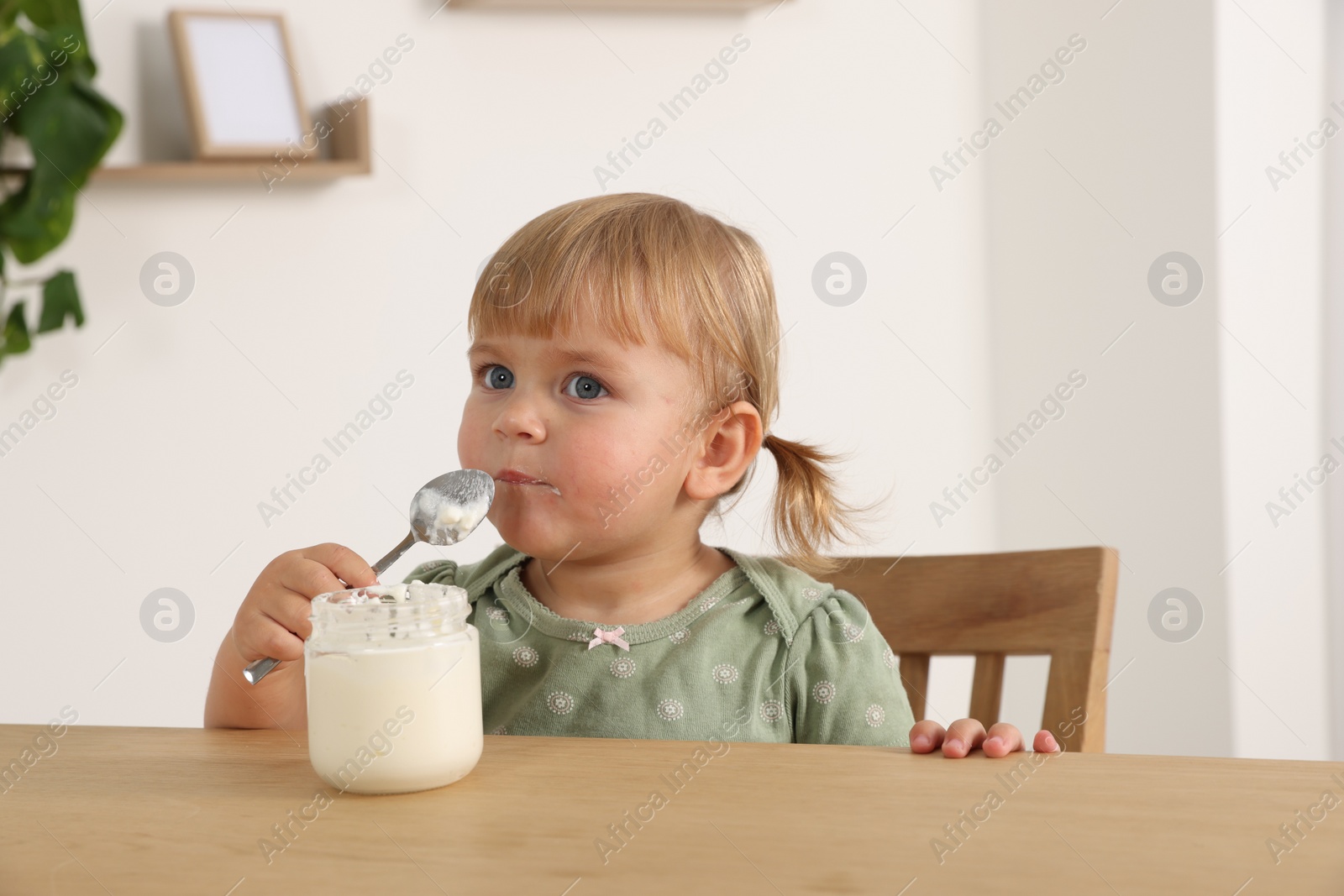 Photo of Cute little child eating tasty yogurt with spoon from jar at wooden table