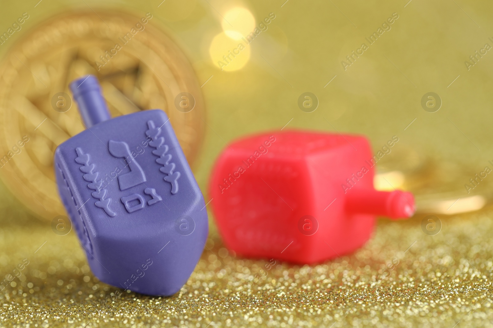 Photo of Hanukkah celebration. Dreidels and coins on golden table against blurred lights, closeup. Space for text