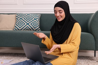 Photo of Muslim woman in hijab using video chat on laptop near sofa indoors