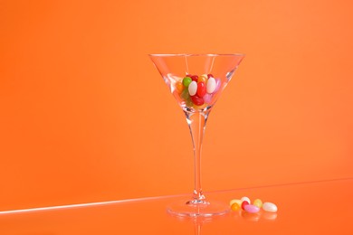 Martini glass with many tasty colorful candies on table against orange background
