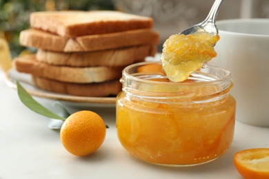 Photo of Spoon with tasty kumquat jam above jar on white table