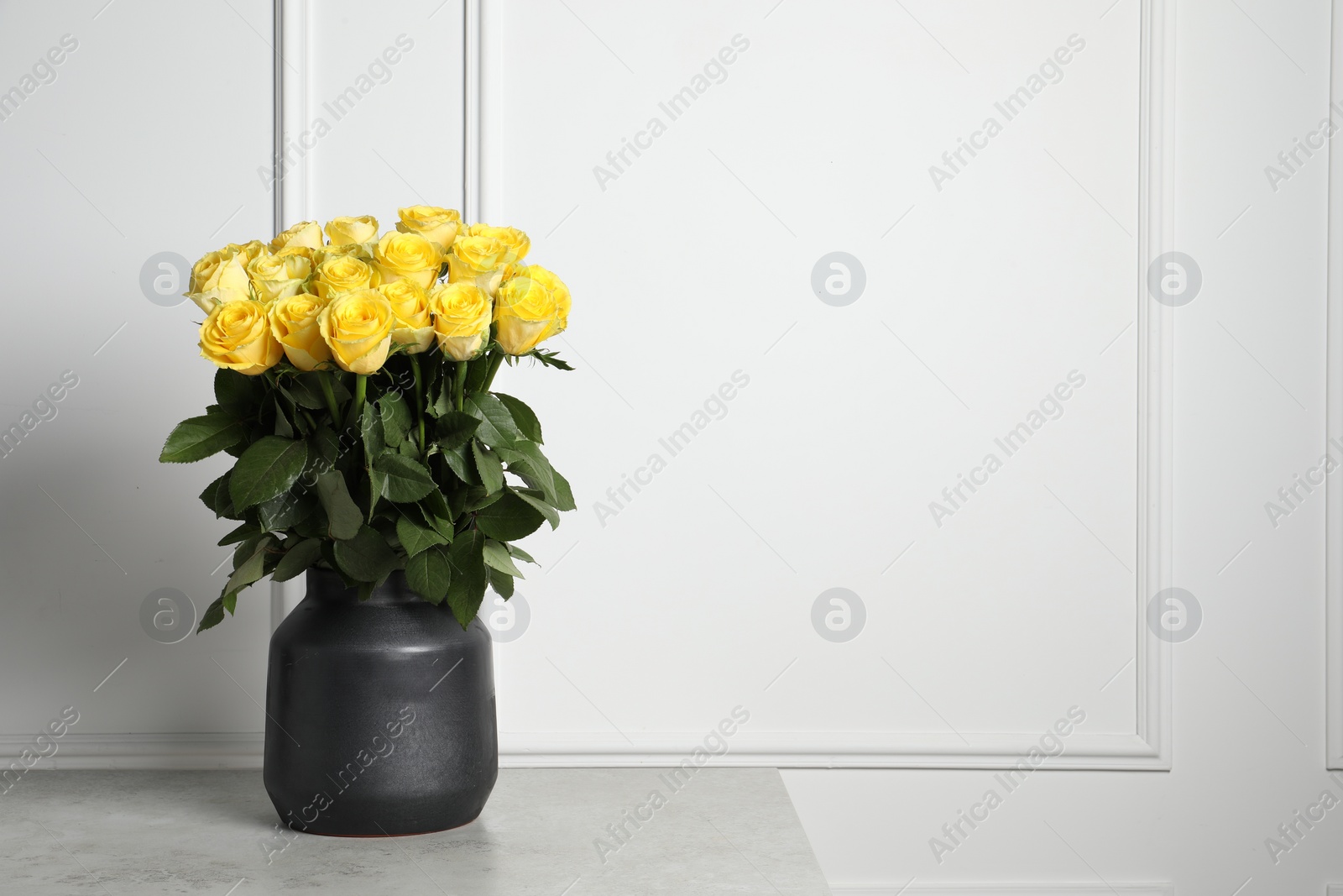 Photo of Beautiful bouquet of yellow roses in vase on light grey table near white wall, space for text