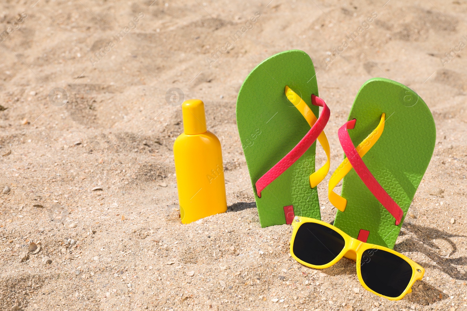 Photo of Stylish flip flops, sunglasses and sun protection cream on sand, space for text