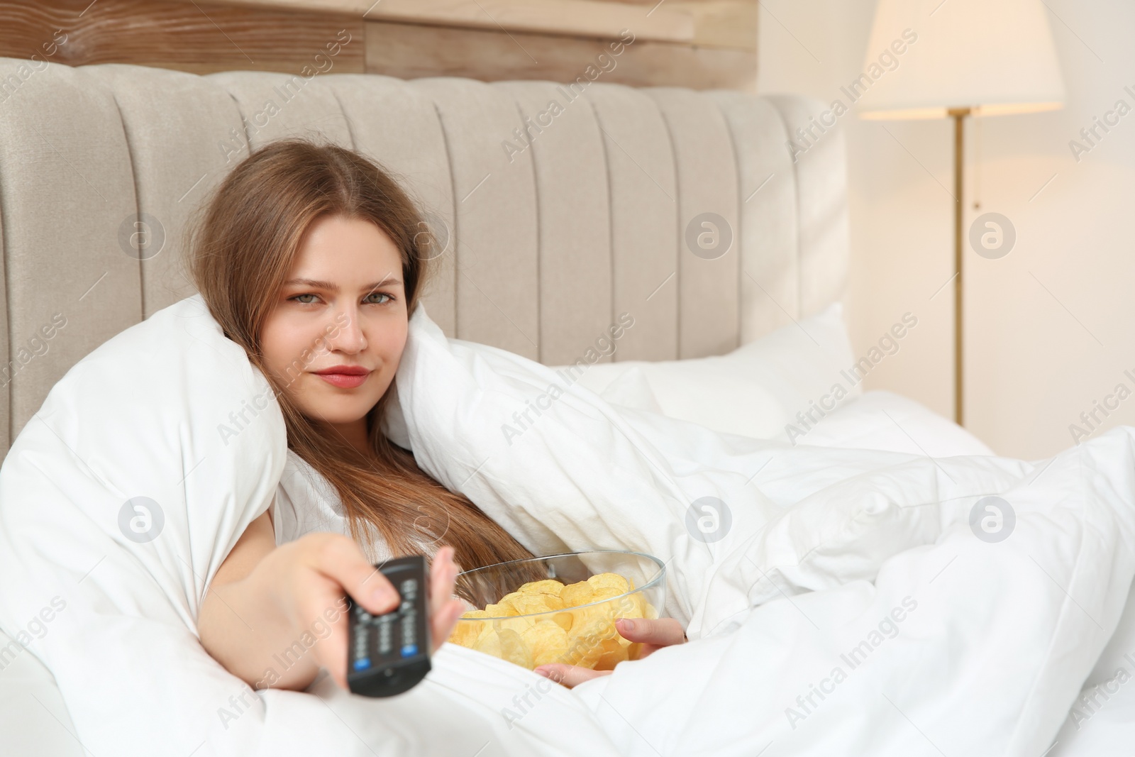 Photo of Lazy young woman with bowl of chips watching TV in bedroom