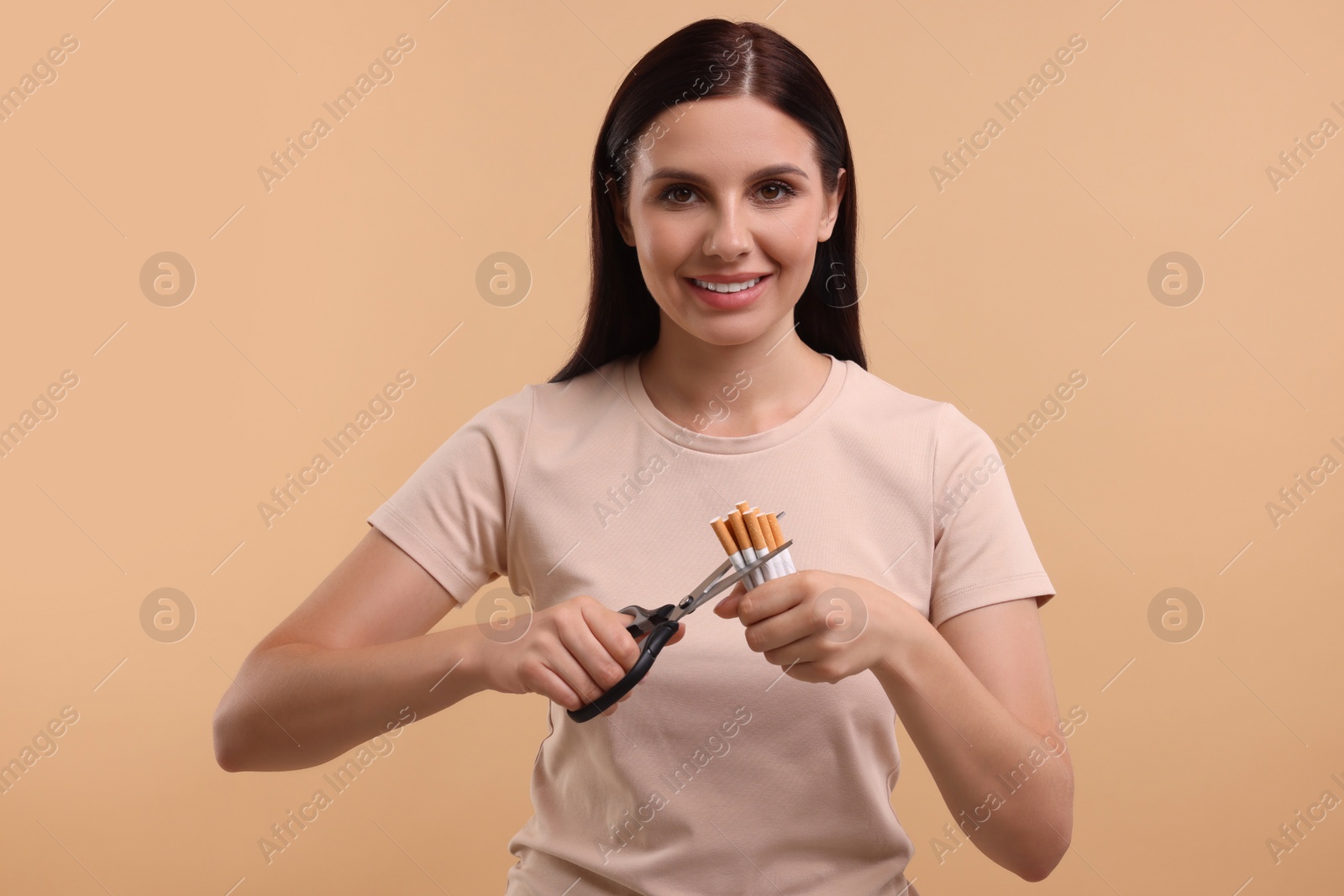 Photo of Stop smoking concept. Happy young woman cutting cigarettes on light brown background