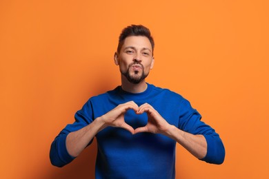 Photo of Handsome man making heart with hands and blowing kiss on orange background