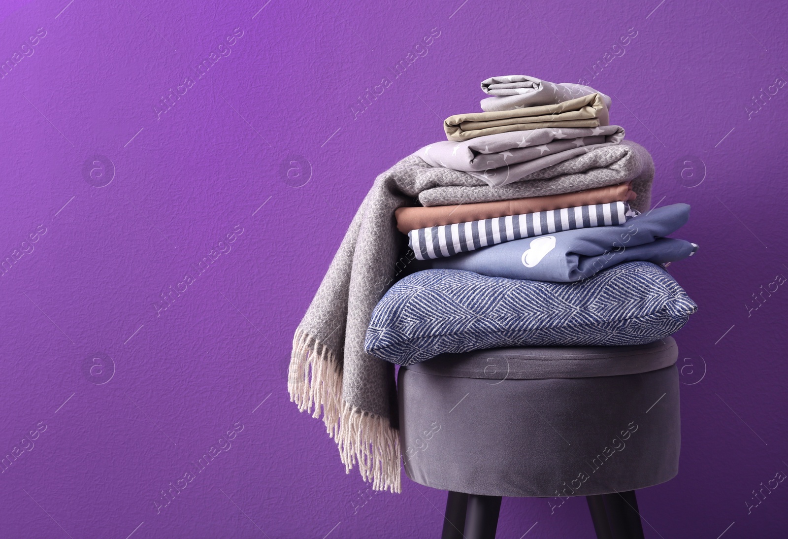 Photo of Stack of clean bed sheets and pillow on stool against purple background. Space for text