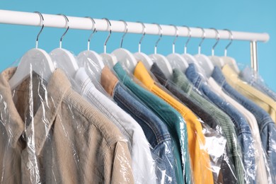 Photo of Dry-cleaning service. Many different clothes in plastic bags hanging on rack against light blue background, closeup