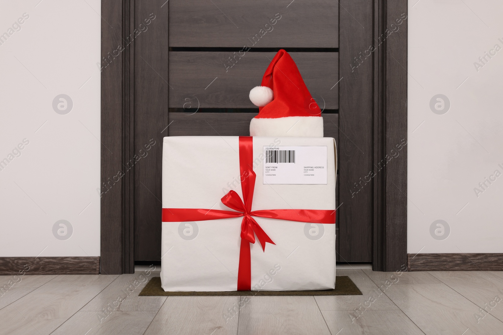 Photo of Christmas gift box and Santa hat near door indoors. Sending present by mail