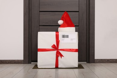 Christmas gift box and Santa hat near door indoors. Sending present by mail