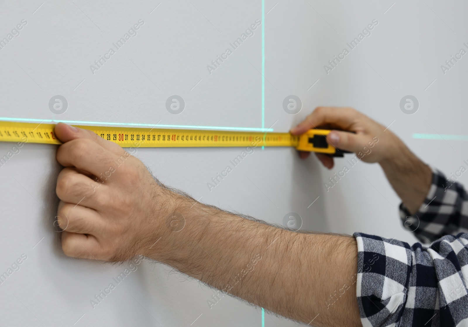 Photo of Man using cross line laser level and tape for accurate measurement on light wall, closeup