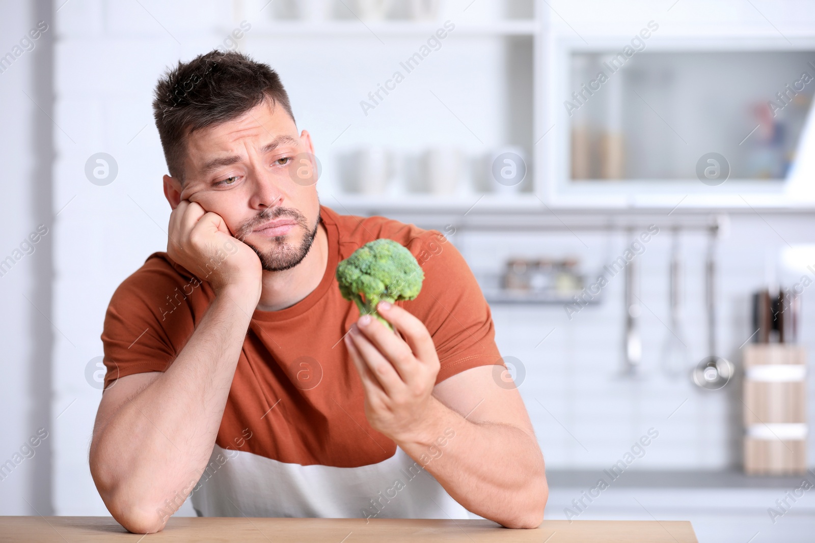 Photo of Portrait of unhappy man with broccoli at table in kitchen