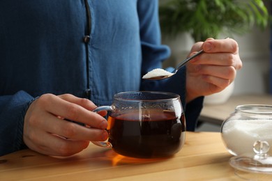 Woman adding sugar into aromatic tea at wooden table indoors, closeup