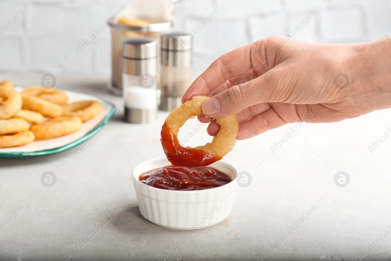 Photo of Woman dipping tasty onion ring into bowl with ketchup on table