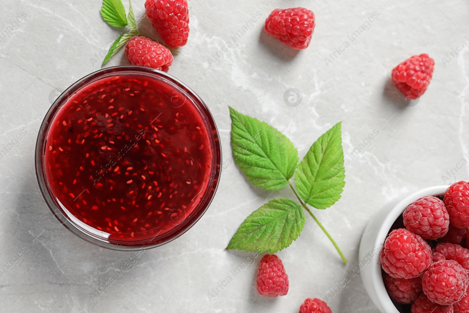 Photo of Bowl of sweet jam with ripe raspberries and green leaves on white table, flat lay