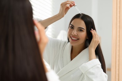 Photo of Beautiful woman applying hair serum indoors, reflection in mirror. Cosmetic product