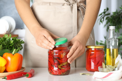 Photo of Woman closing jar with pickled peppers at white table, closeup