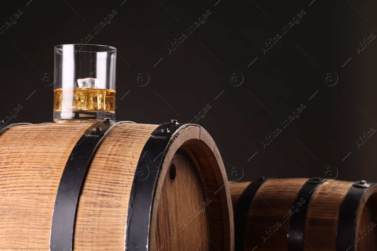 Photo of Whiskey with ice cubes in glass on wooden barrel against dark background, space for text