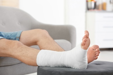 Photo of Young man with broken leg in cast sitting on sofa at home