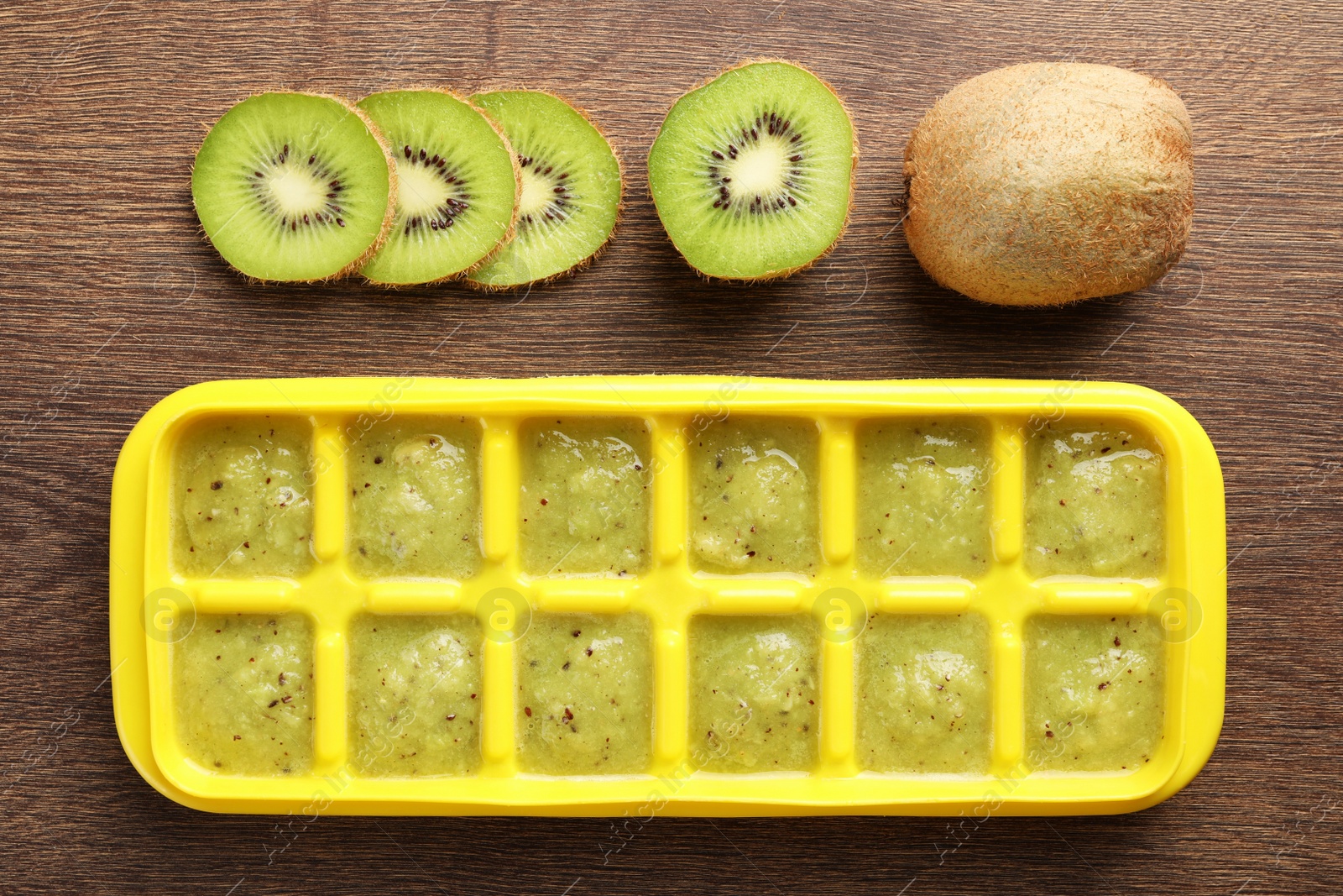 Photo of Kiwi puree in ice cube tray and fresh kiwi fruits on wooden table, flat lay. Ready for freezing