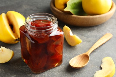 Tasty homemade quince jam in jar, spoon and fruits on grey textured table, closeup