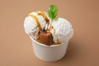 Scoops of tasty ice cream with caramel sauce, mint leaves and candy in paper cup on light brown table, closeup