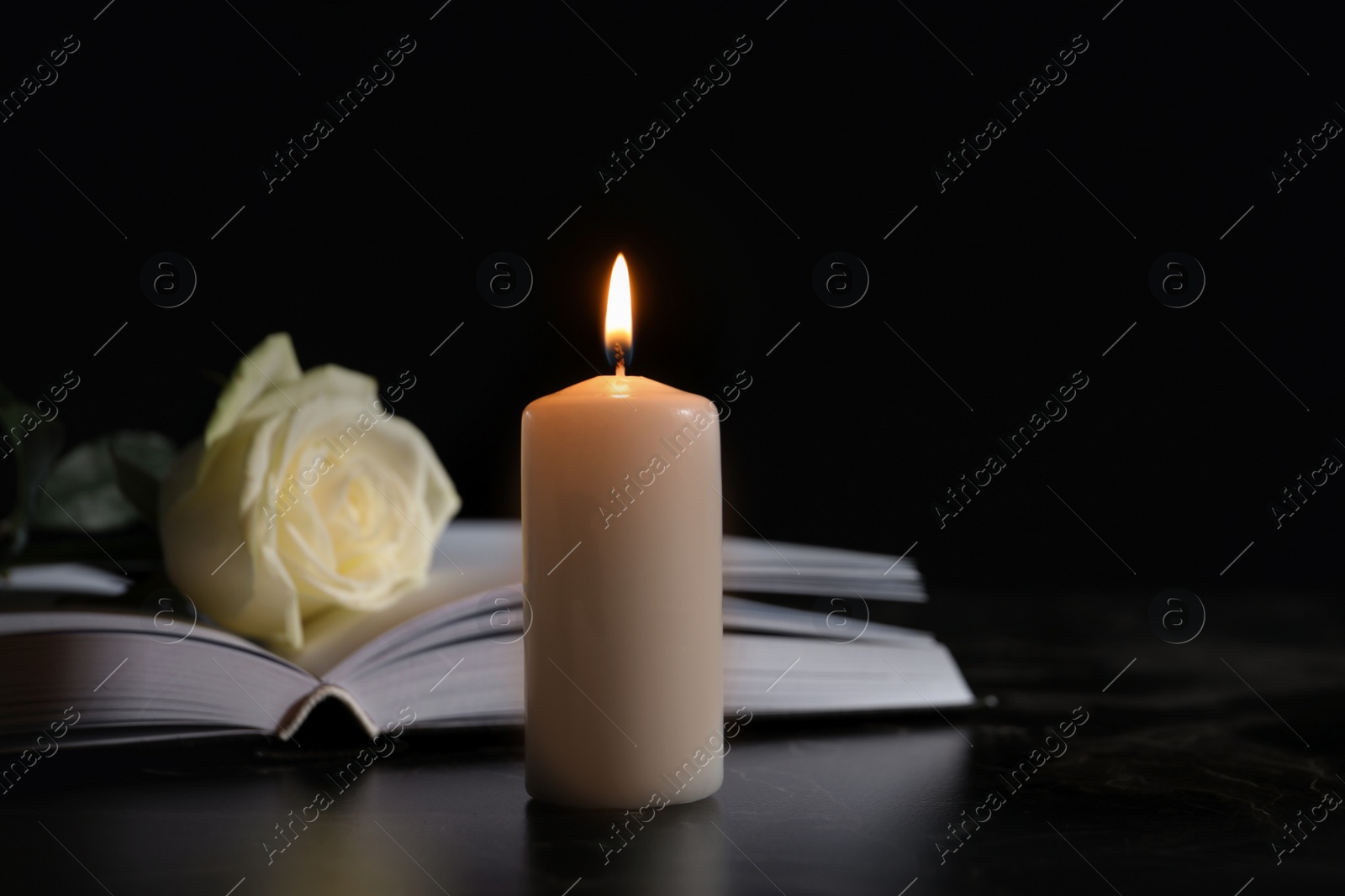 Photo of Burning candle, book and white rose on table in darkness, space for text. Funeral symbol