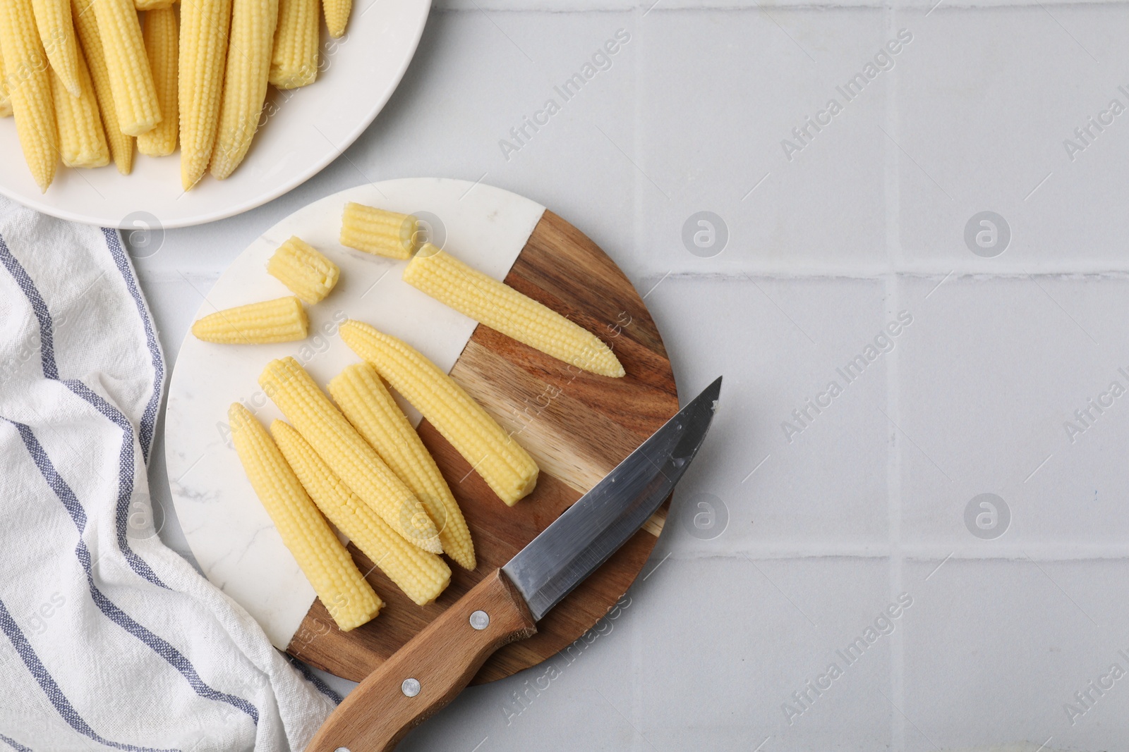 Photo of Tasty fresh yellow baby corns and knife on white tiled table, top view. Space for text