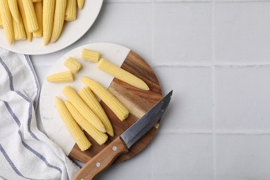 Tasty fresh yellow baby corns and knife on white tiled table, top view. Space for text