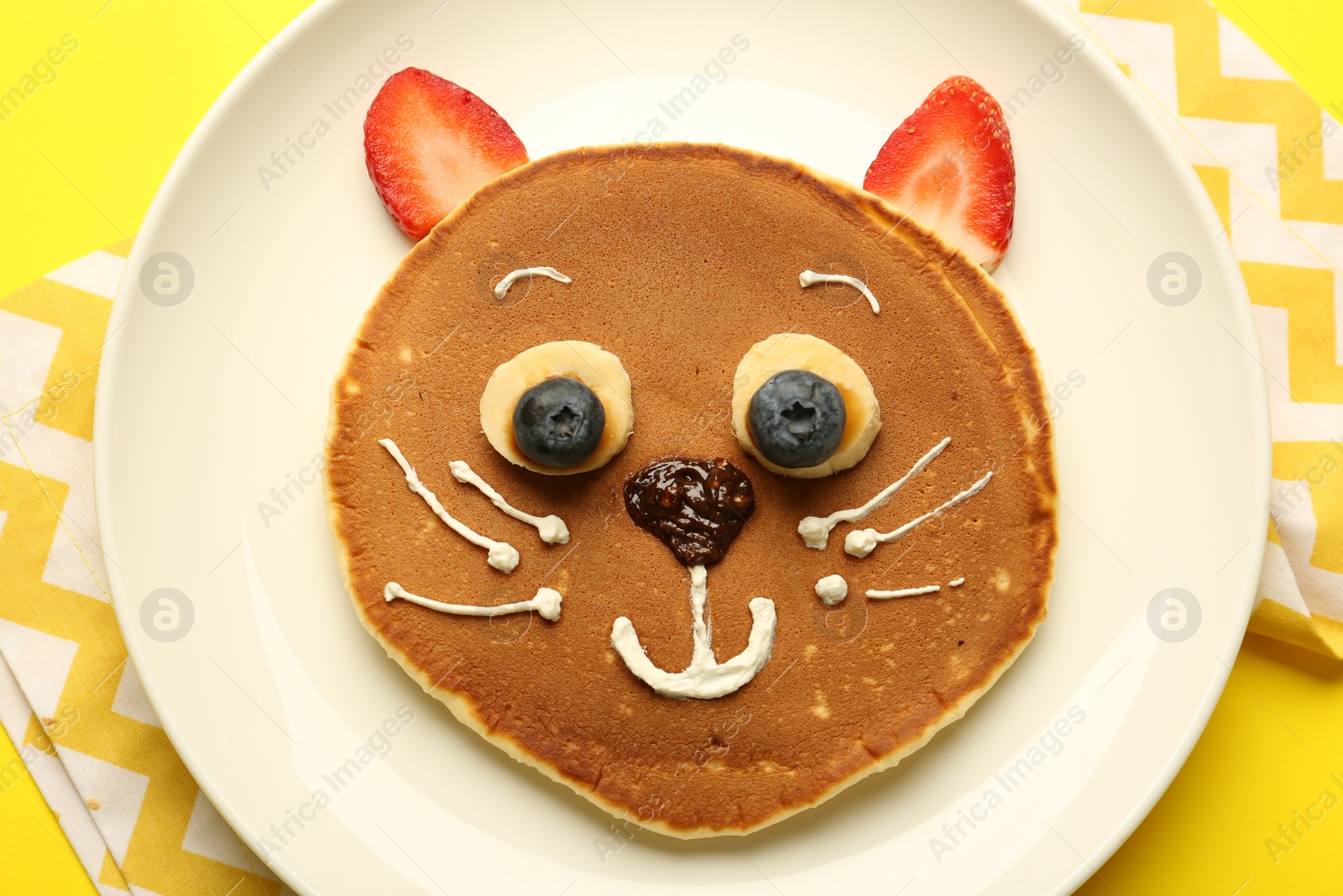 Photo of Creative serving for kids. Plate with cute cat made of pancakes, berries, cream, banana and chocolate paste on yellow background, top view