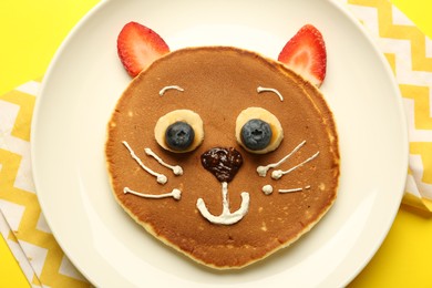 Creative serving for kids. Plate with cute cat made of pancakes, berries, cream, banana and chocolate paste on yellow background, top view