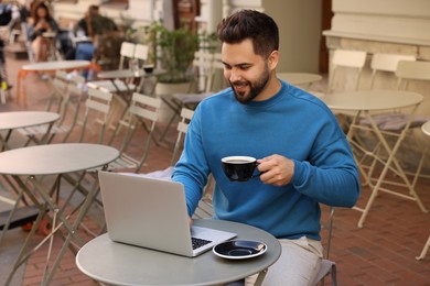 Handsome young man with cup of drink working on laptop at table in outdoor cafe