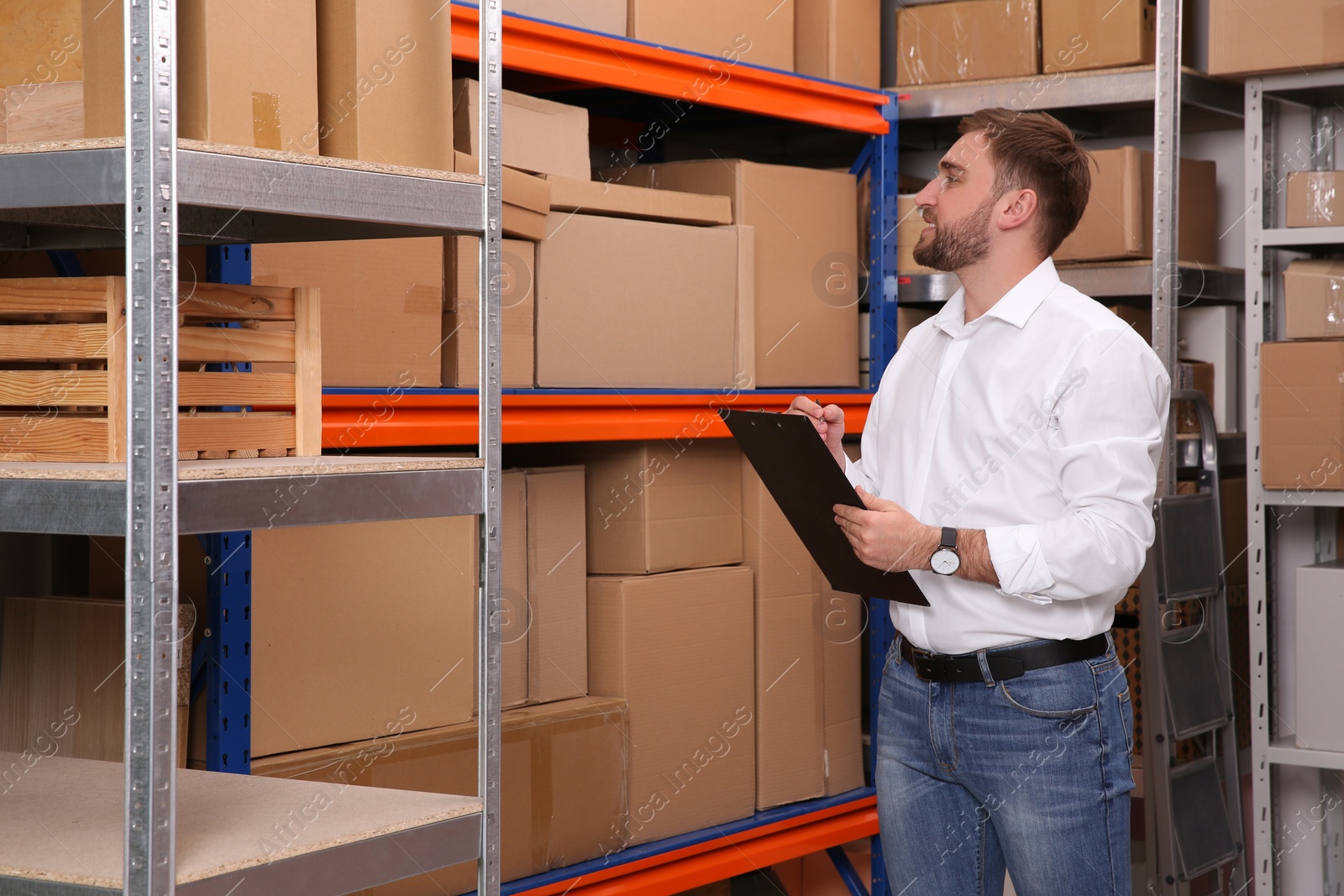 Photo of Young businessman with clipboard near rack of cardboard boxes at warehouse