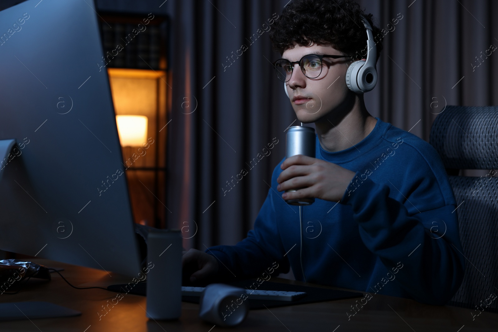 Photo of Young man with energy drink and headphones playing video game at wooden desk indoors