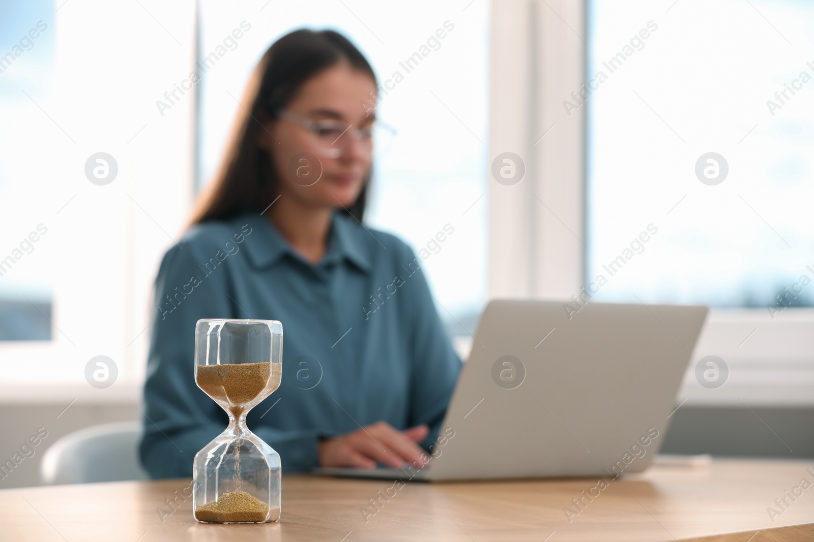 Photo of Hourglass with flowing sand on table. Woman using laptop indoors, selective focus