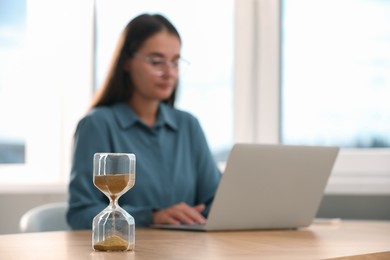 Hourglass with flowing sand on table. Woman using laptop indoors, selective focus