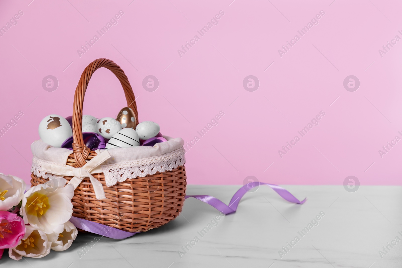 Photo of Wicker basket with festively decorated Easter eggs and beautiful tulips on white marble table against pink background. Space for text