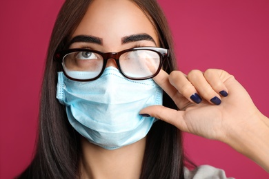 Photo of Young woman wiping foggy glasses caused by wearing disposable mask on pink background. Protective measure during coronavirus pandemic