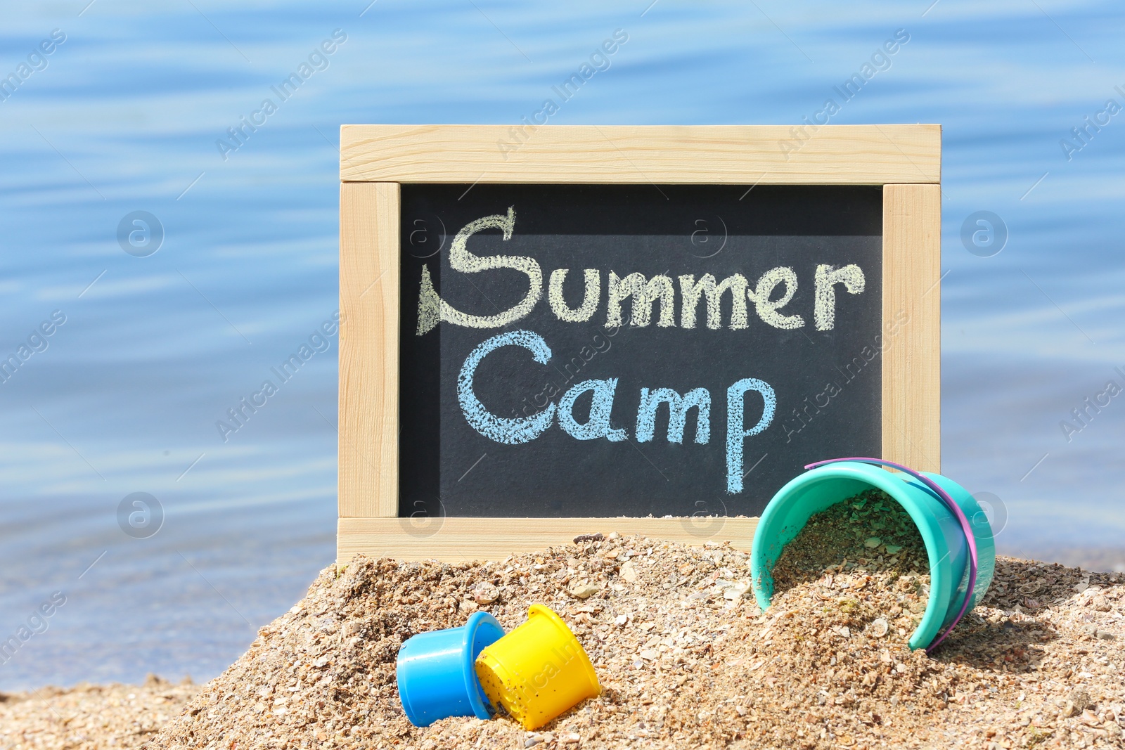 Photo of Little blackboard with text SUMMER CAMP and toys on sand near sea