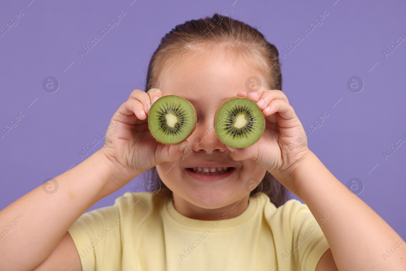 Photo of Smiling girl covering eyes with halves of fresh kiwi on violet background