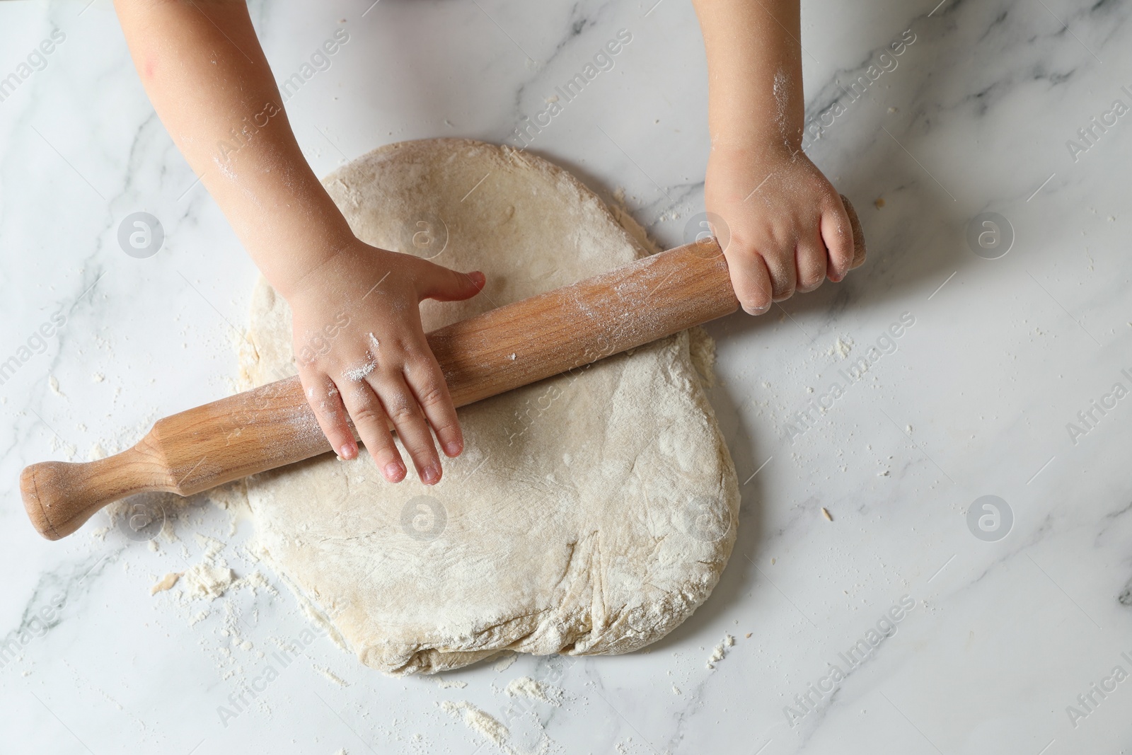 Photo of Little child rolling raw dough at white table, top view