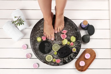 Photo of Woman soaking her feet in plate with water, stones, flowers and lime slices on white wooden floor, top view. Pedicure procedure