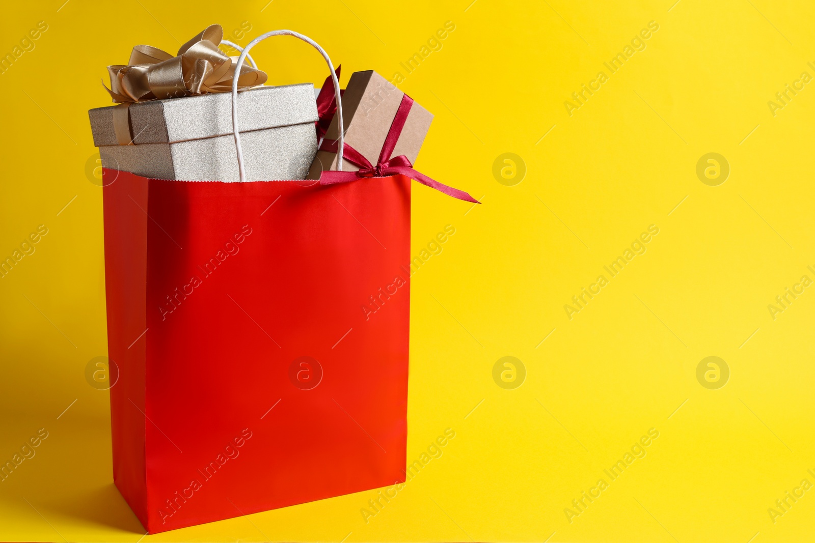 Photo of Red paper shopping bag full of gift boxes on yellow background. Space for text