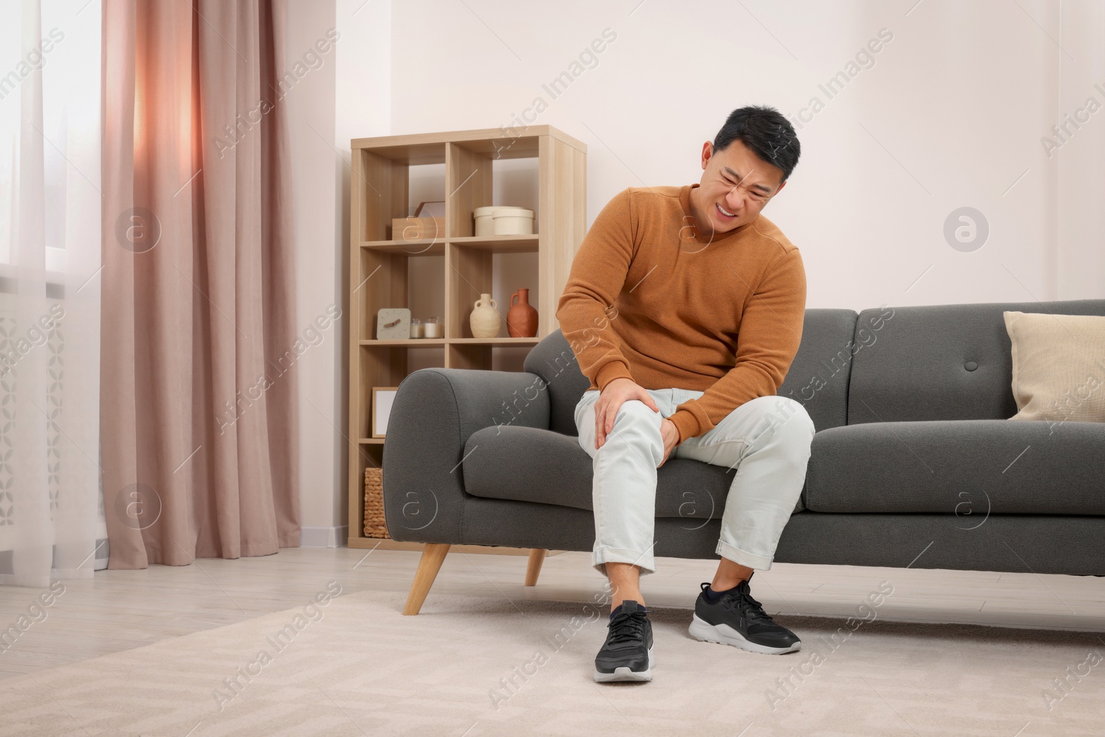 Photo of Asian man suffering from knee pain on sofa indoors