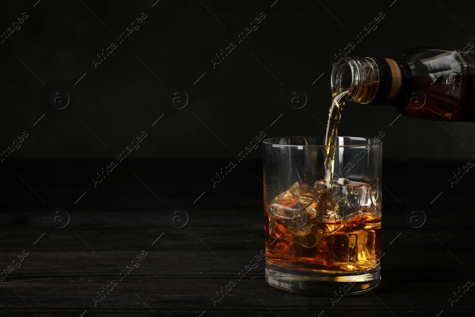 Photo of Pouring whiskey from bottle into glass with ice cubes on table. Space for text